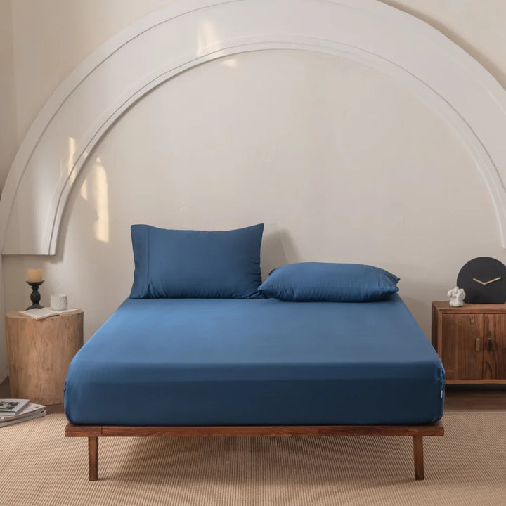 A minimalist bedroom highlights a wooden bed frame with luxurious blue bedding and Linenly's Bamboo Fitted Sheet in Midnight for extra softness. The room features softly neutral arched walls, a wooden nightstand with a clock, and a candle on a stool. Natural light creates gentle shadows.