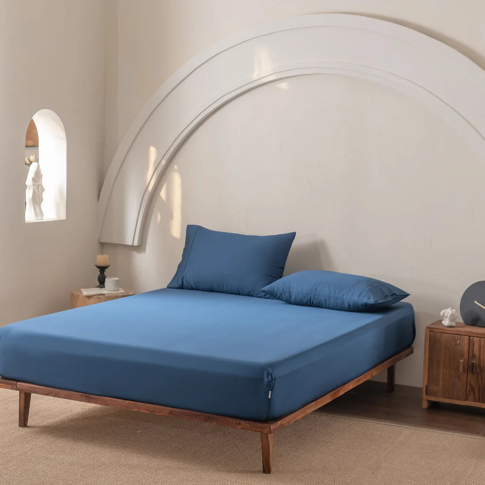 A minimalist bedroom showcases a wooden bed frame with Linenly’s Bamboo Fitted Sheet Set in Midnight. An arched wall detail adorns above, while a wooden side table features a candle and decor. Soft, natural light from an arched window on the left enhances the serene atmosphere.