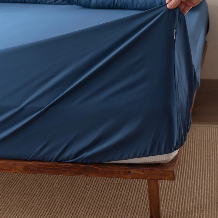 A hand gently holds a corner of the Linenly Bamboo Fitted Sheet - Midnight, draping its luxurious softness over the mattress on an elegant wooden bed frame with a woven beige rug beneath.