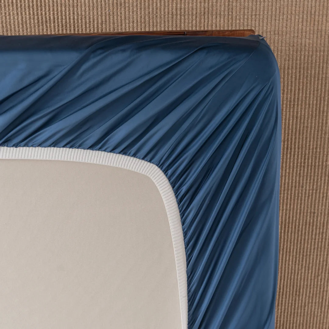 A corner of a mattress covered by a Linenly Bamboo Fitted Sheet - Midnight in blue, shows its white border. Beneath is a textured surface, possibly neutral-toned carpet or rug, enhancing the feel of softness and luxury.