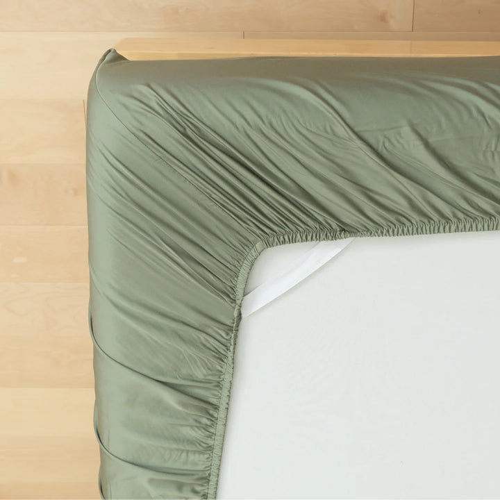 A neatly made bed with a Linenly Bamboo Fitted Sheet in Moss, a smooth and breathable fabric, on the mattress against a wooden headboard background.