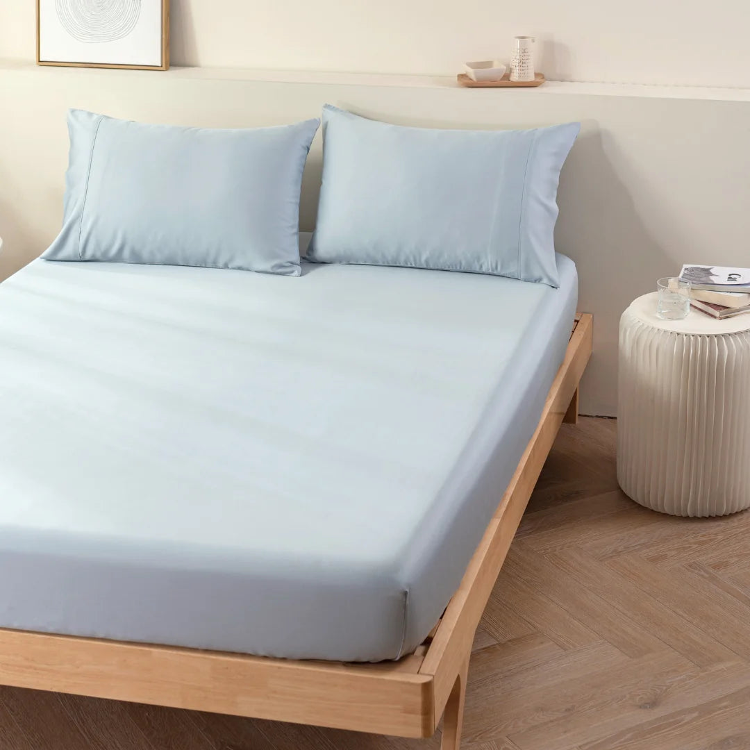 A minimalistic bedroom with a wooden bed frame featuring neatly arranged Linenly Bamboo Fitted Sheet Set in pale blue and matching pillows. Beside the bed is a small, white round bedside table adorned with decorative items and a cup. The room exudes an eco-friendly, light, and airy ambiance with a cooling feel.