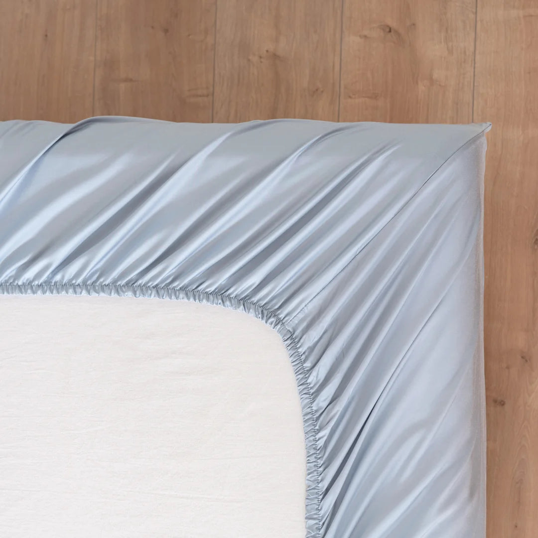 A neatly placed pale blue-colored bamboo fitted sheet by Linenly on a mattress, exhibiting a smooth and silky texture with a cooling touch, with a wooden floor in the background.