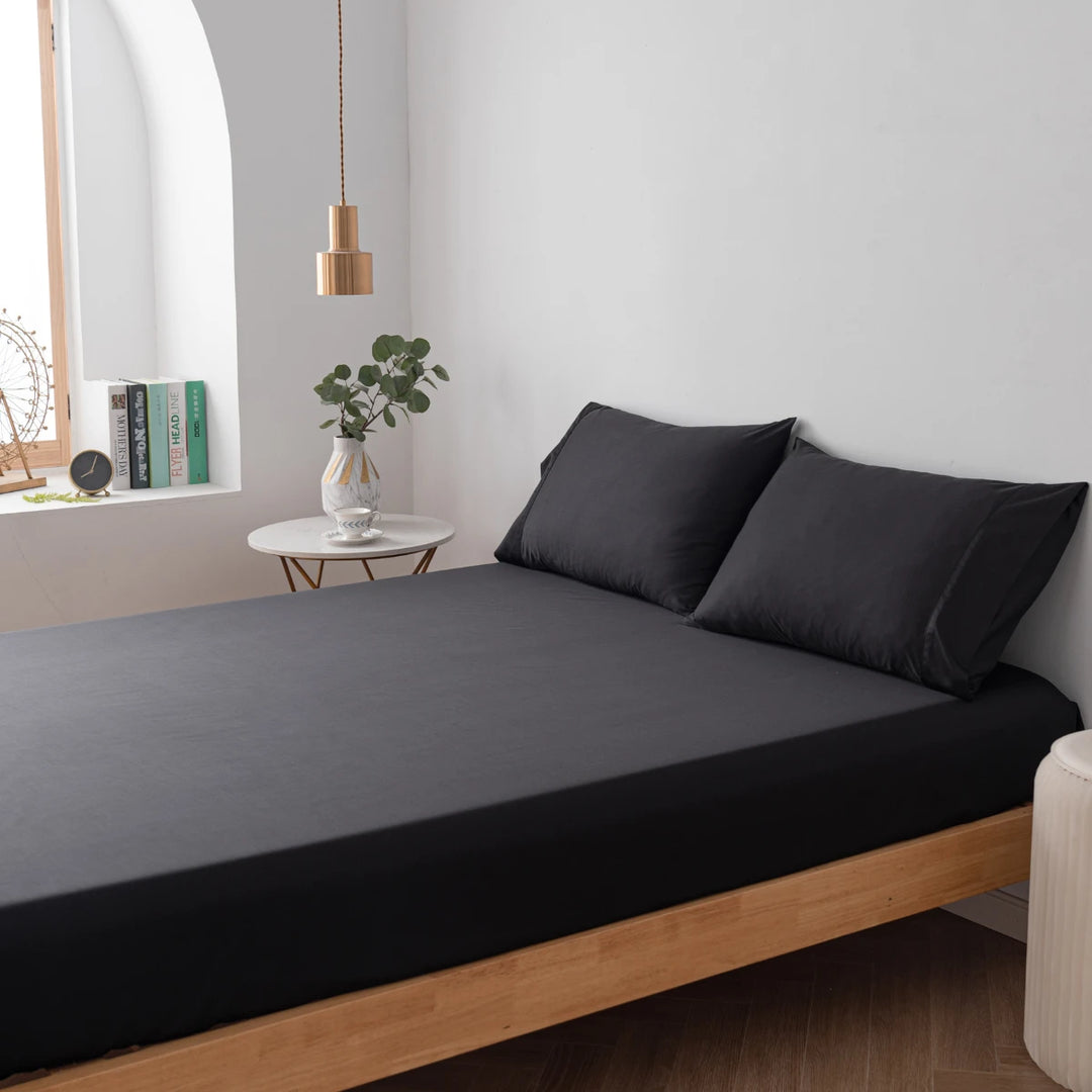 A modern minimalist bedroom with a low bed frame, Linenly Black Bamboo Fitted Sheets, a small side table with a plant, and a hanging light fixture.