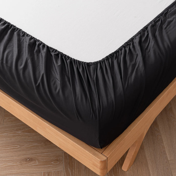 A luxurious Linenly black bamboo fitted sheet on a bed with a wooden frame, neatly placed over a light wood floor.