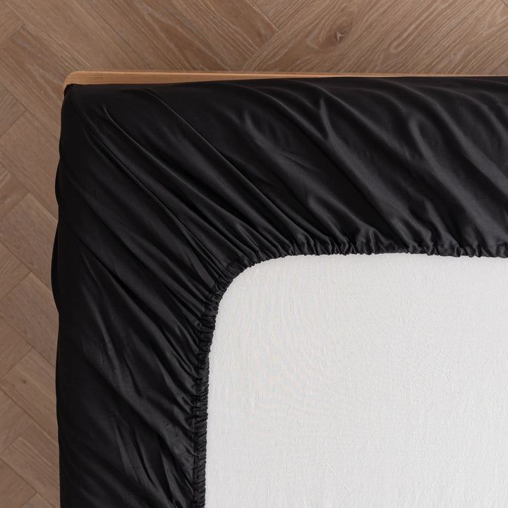 A corner of a neatly made bed with Linenly's black Bamboo Fitted Sheet, offering unparalleled comfort on a light-colored mattress, against a wooden floor background.