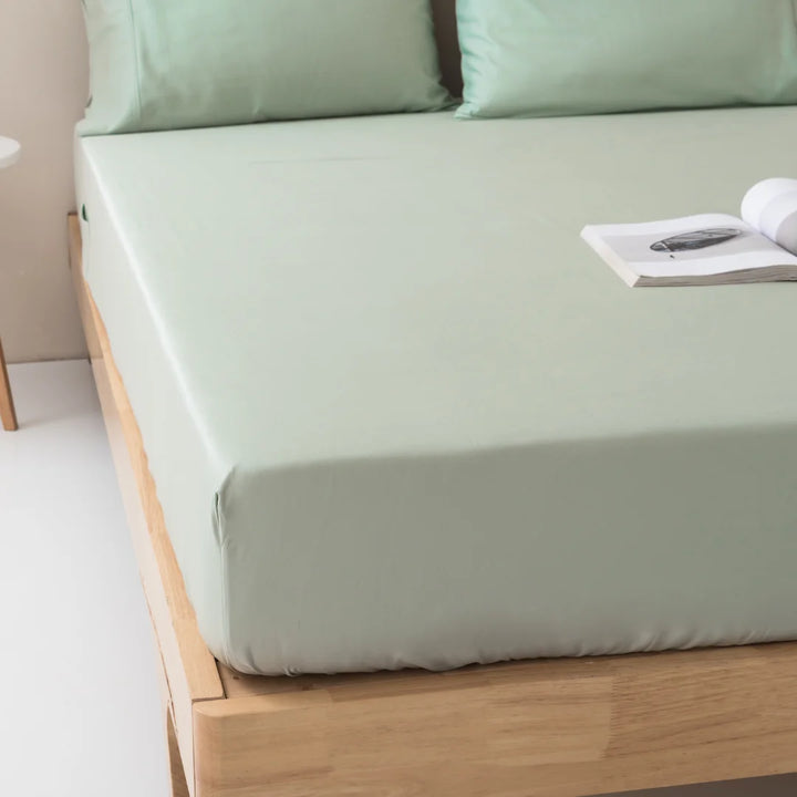 A neatly made bed with a smooth, pastel green Linenly Bamboo Fitted Sheet in Sage, complemented by matching pillows in a serene and minimalist bedroom setting. A magazine rests on the bed, adding a touch.