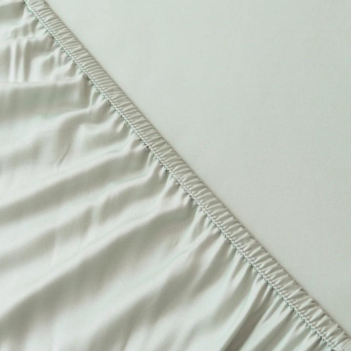 A close-up view of a neatly-stitched seam where two pieces of white, eco-friendly Linenly bamboo fitted sheet in sage meet, with one side featuring a smooth texture and the other exhibiting a slight sheen and softness.