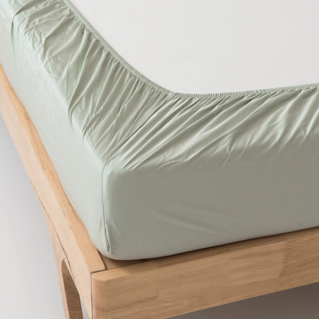Sentence with replacement: A light green Linenly bamboo fitted bedsheet in Sage neatly wrapped around the corner of a wooden bed frame, highlighting a smooth and wrinkle-free texture.
