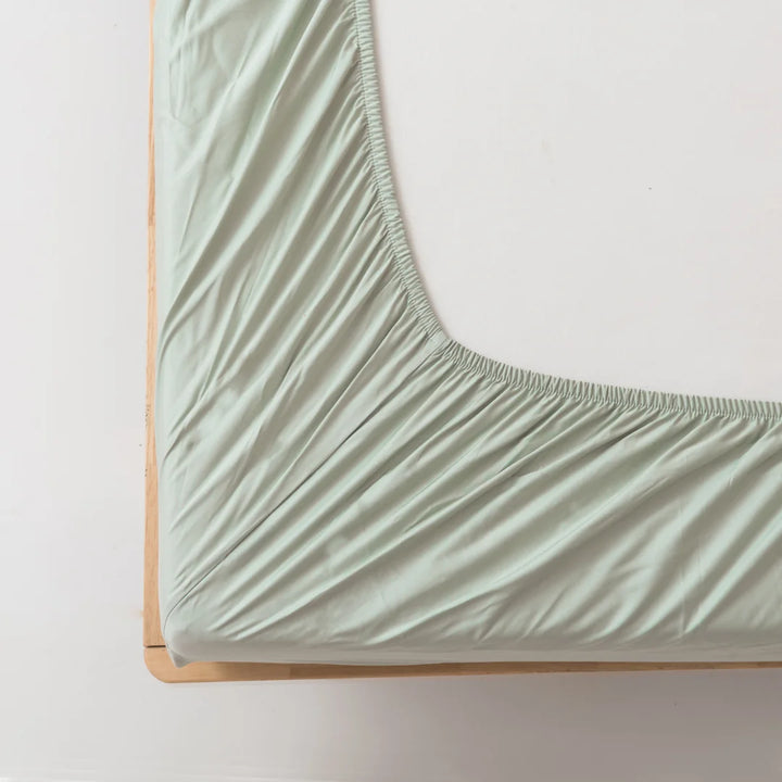 A top-down view of a neatly made bed with soft green, eco-friendly Linenly Bamboo Fitted Sheet in Sage exhibiting gentle creases, tightly fitted over a mattress on a simple bed frame, against a white background.