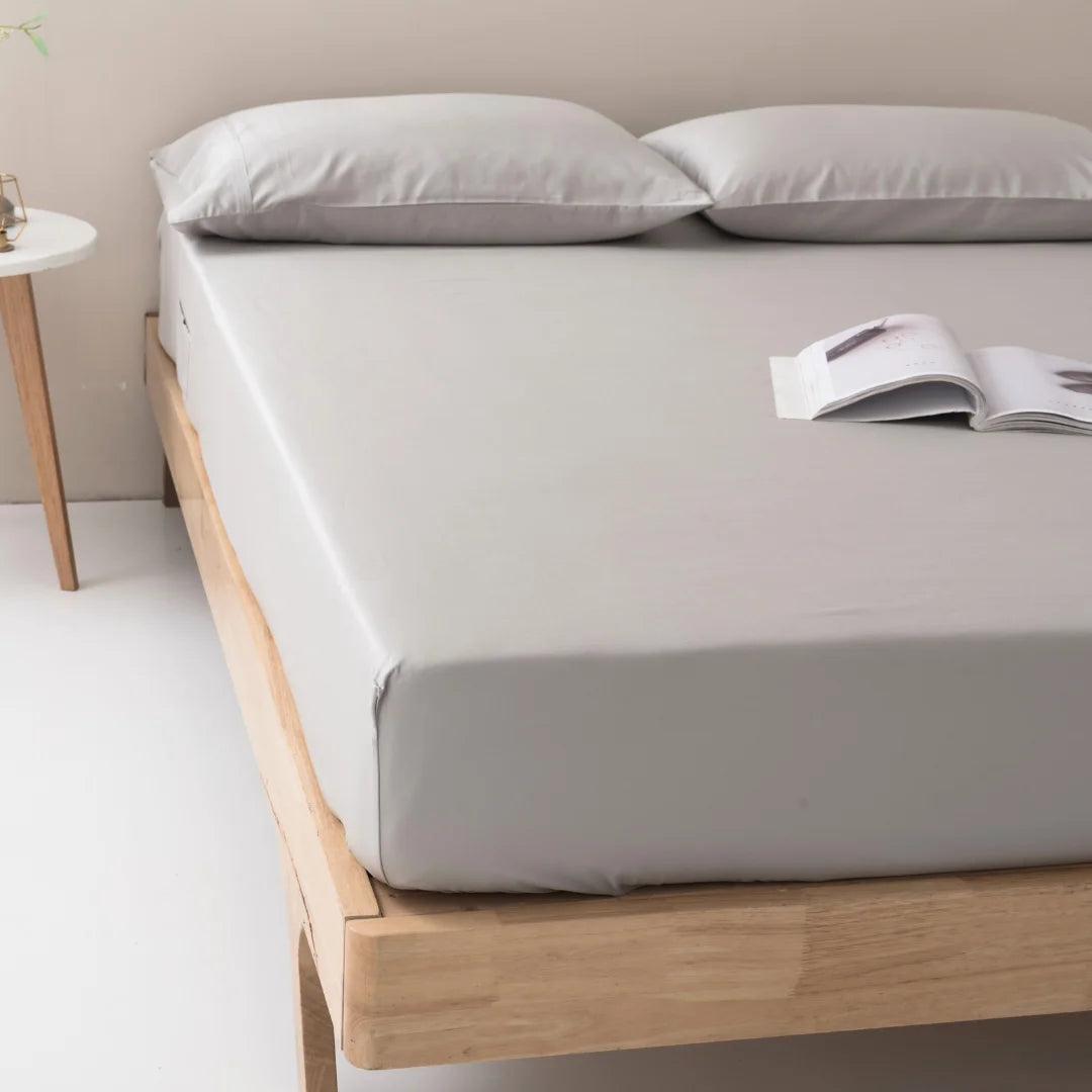 A neatly made bed with Linenly's light gray Bamboo Fitted Sheet - Silver and a pair of fluffy pillows, with an open magazine resting at the foot of the bed, beside a simple wooden nightstand.