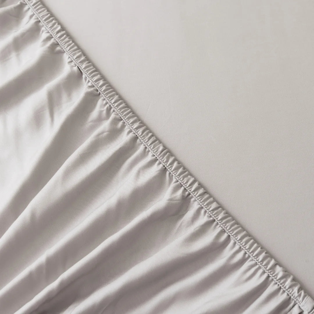 A close-up image showcases the detailed stitching between two pieces of sustainable luxury fabric, with one part crafted from Linenly's Bamboo Fitted Sheet - Silver appearing silky and reflecting light, while the other remains matte and smooth.