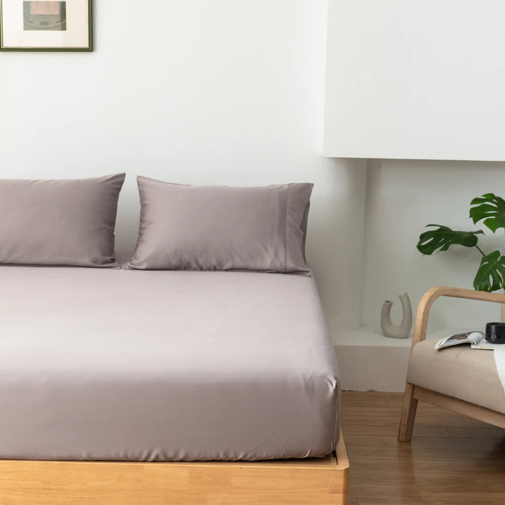 A neatly made bed with eco-friendly Linenly Bamboo Fitted Sheet in a minimalist bedroom setting, accompanied by a side table with a book and a green houseplant.