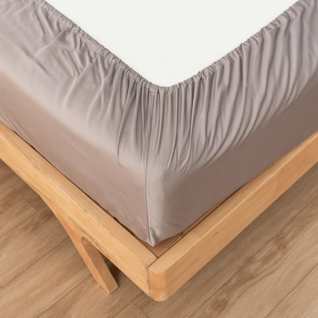 A neatly fitted grey Linenly Bamboo Fitted Sheet - Stone Terrace on a wooden bed frame, with the mattress on top and a white mattress protector visible, set against a hardwood floor background.