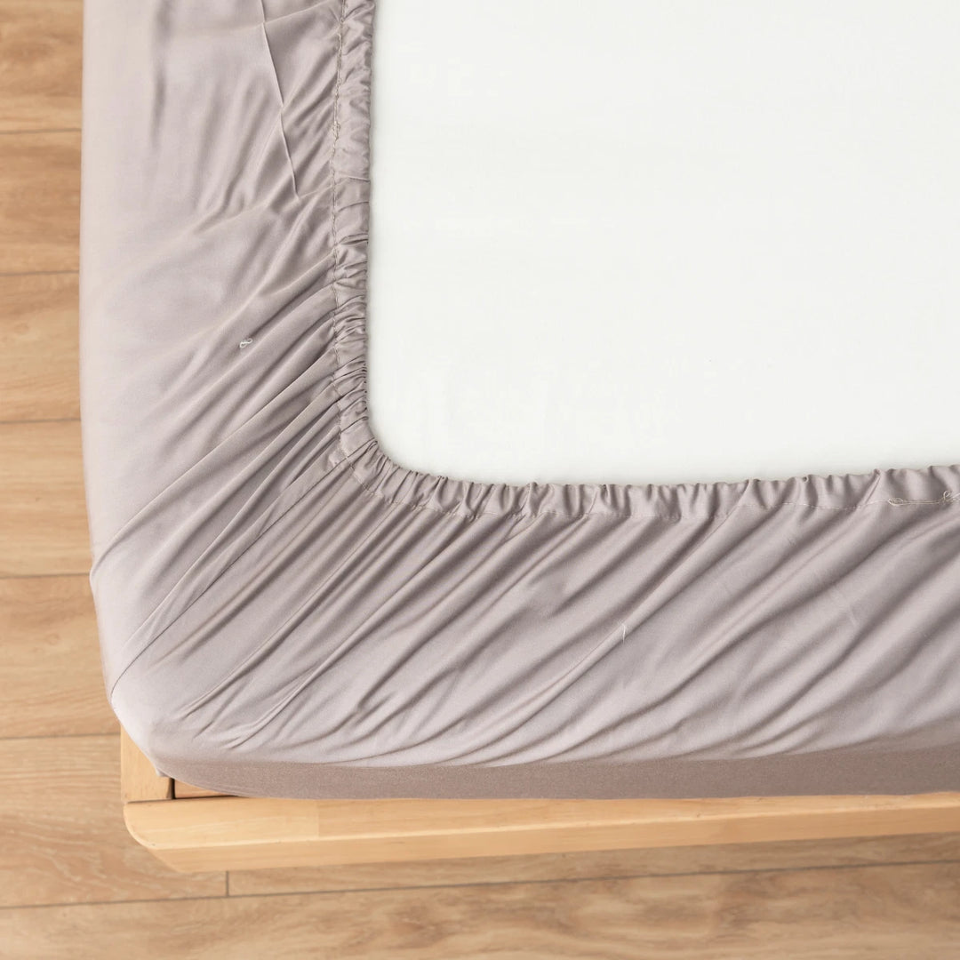 A neatly fitted grey Bamboo Fitted Sheet - Stone Terrace on a mattress with an elasticated corner, set on a simple wooden bed frame by Linenly.