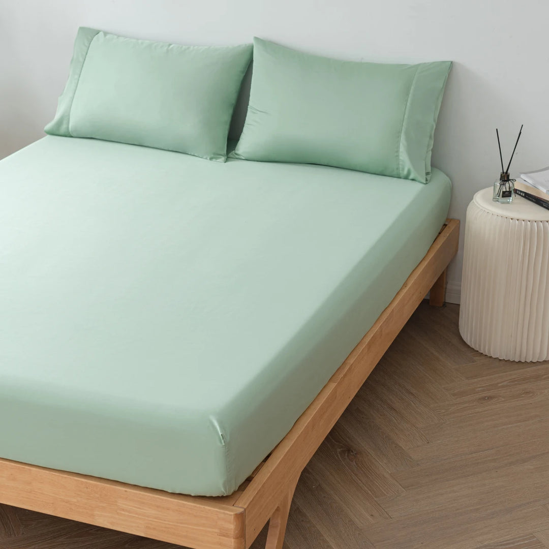 A neatly made bed with eco-friendly, Linenly Summer Green bamboo fitted sheets on a wooden bed frame, accompanied by a matching pillowcase set, beside a white bedside table with decorative items on it.