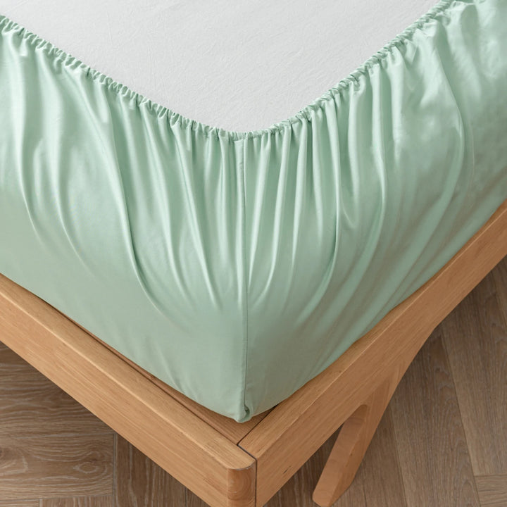 A neatly made bed with Linenly's Summer Green Bamboo Fitted Sheet on a wooden bed frame, against a simple background with a light-colored floor.