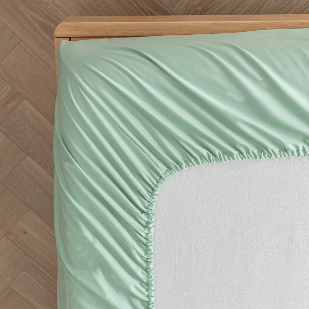 A neatly made eco-friendly bed with Linenly Summer Green bamboo fitted sheets on a wooden frame, showcased in a room with herringbone parquet flooring.