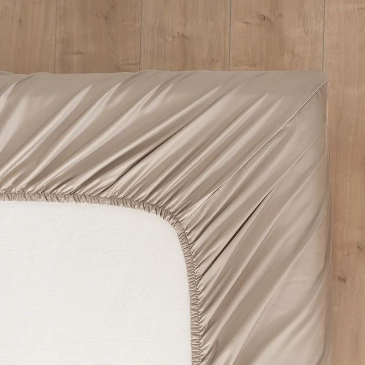 An elegantly made bed with a smooth, Linenly taupe bamboo fitted sheet and a neatly tucked corner showcasing luxury bedding on a wooden floor, promising a breathable bamboo fabric experience that transforms your space into a luxury.