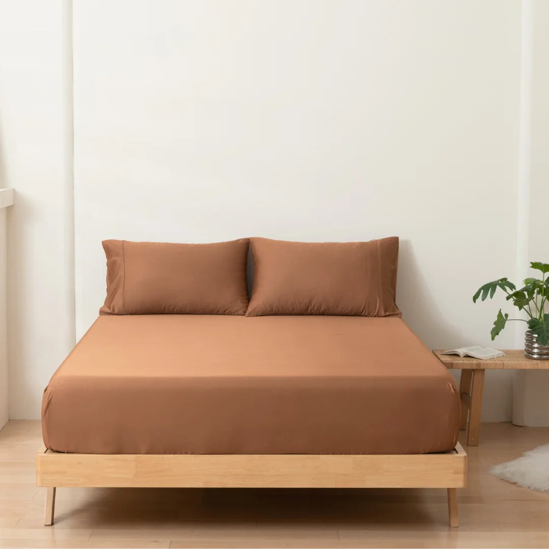 A neatly made bed with Linenly terracotta bamboo fitted sheets in a minimalist bedroom interior, featuring a wooden bed frame, a small bedside table, and a green potted plant adding a touch of nature.