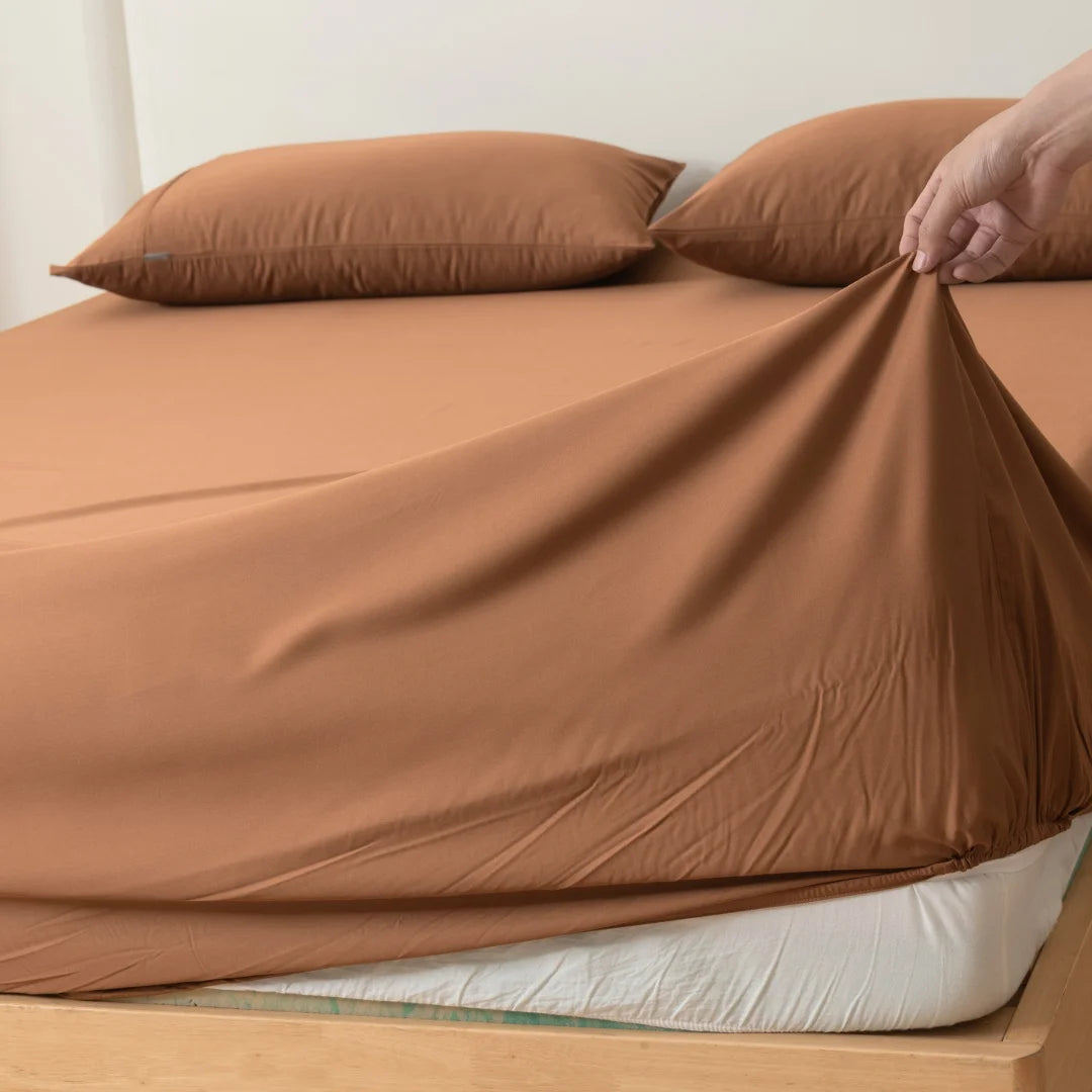 A person is neatly tucking in luxurious Linenly Bamboo Fitted Sheets in Terracotta on a bed, ensuring a smooth and snug fit over the mattress.