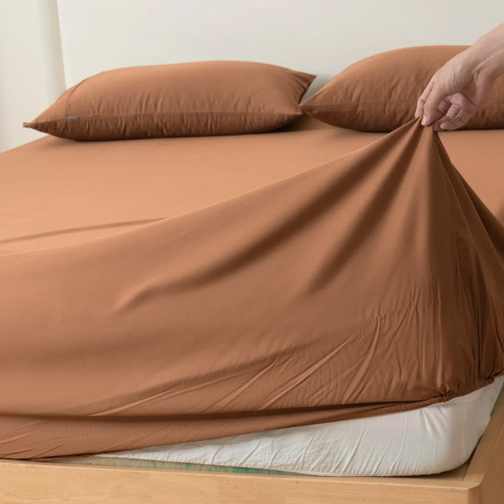 A person is neatly tucking in luxurious Linenly Bamboo Fitted Sheets in Terracotta on a bed, ensuring a smooth and snug fit over the mattress.
