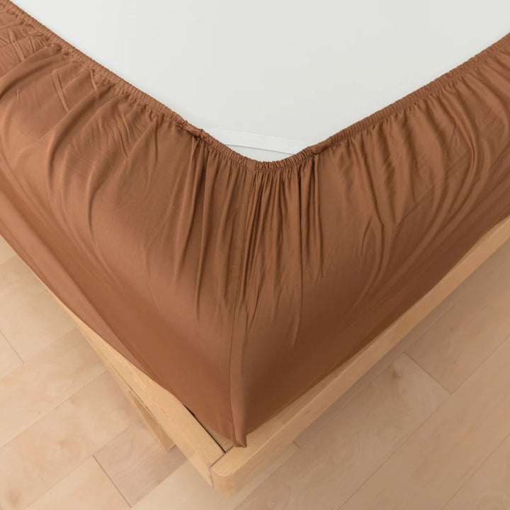 A neatly arranged chocolate brown bed skirt on a bed, complemented by luxurious Linenly Terracotta Bamboo Fitted Sheets, with a smooth wooden floor in the background.
