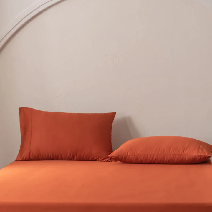 A neatly made bed showcases the Linenly Bamboo Pillowcase Set in Copper Sunset. One pillow lies flat on the right, while another stands upright on the left. The pale, curved wall behind enhances the minimalist aesthetic for a cozy and relaxing atmosphere.