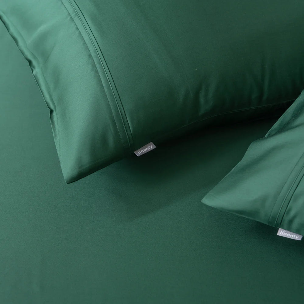 Close-up of a bed featuring Linenly's dark green satin weave sheets and the Bamboo Pillowcase Set in Evergreen. The smooth, well-crafted fabric and visible label enhance a cozy, elegant atmosphere.