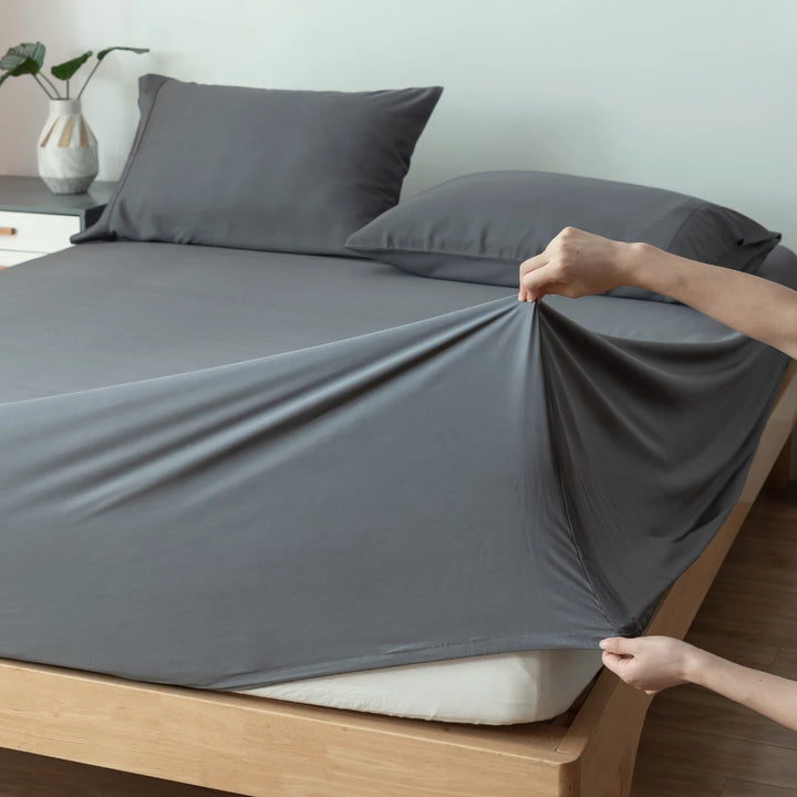 A person making the bed, neatly tucking in a gray fitted sheet on a mattress, complemented with Linenly's Bamboo Pillowcase Set in Granite Grey.