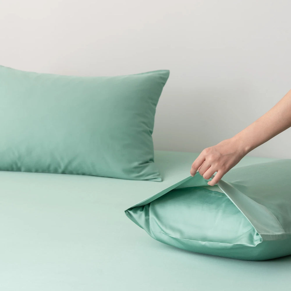 A person's hand gently lifting the corner of a soft, light green pillow with Linenly Bamboo Pillowcase Set - Green Sheen on a matching green background, evoking a calm and cohesive aesthetic.