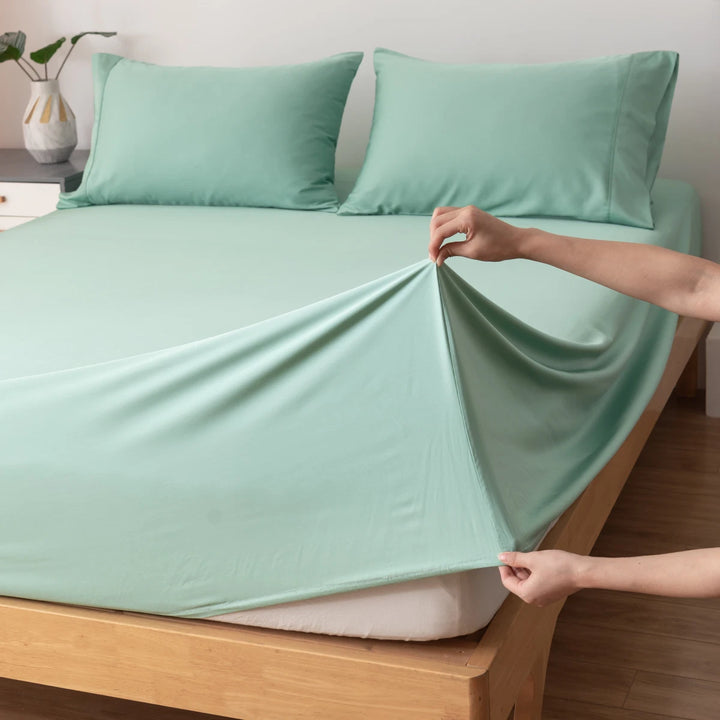 A person's hands pulling a Linenly Bamboo Pillowcase Set - Green Sheen tight over the corner of a neatly made bed, with matching satin weave pillows in the background.