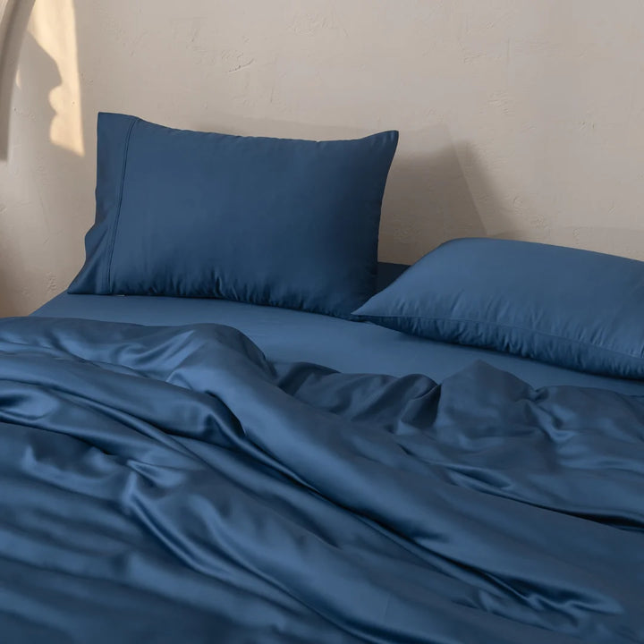 Blue bedding featuring Linenly's Bamboo Pillowcase Set - Midnight and a blanket is neatly arranged on a bed. Soft lighting casts subtle shadows, creating a cozy and inviting atmosphere against the textured cream-colored wall, highlighting the satin weave of the organic bamboo fabric.