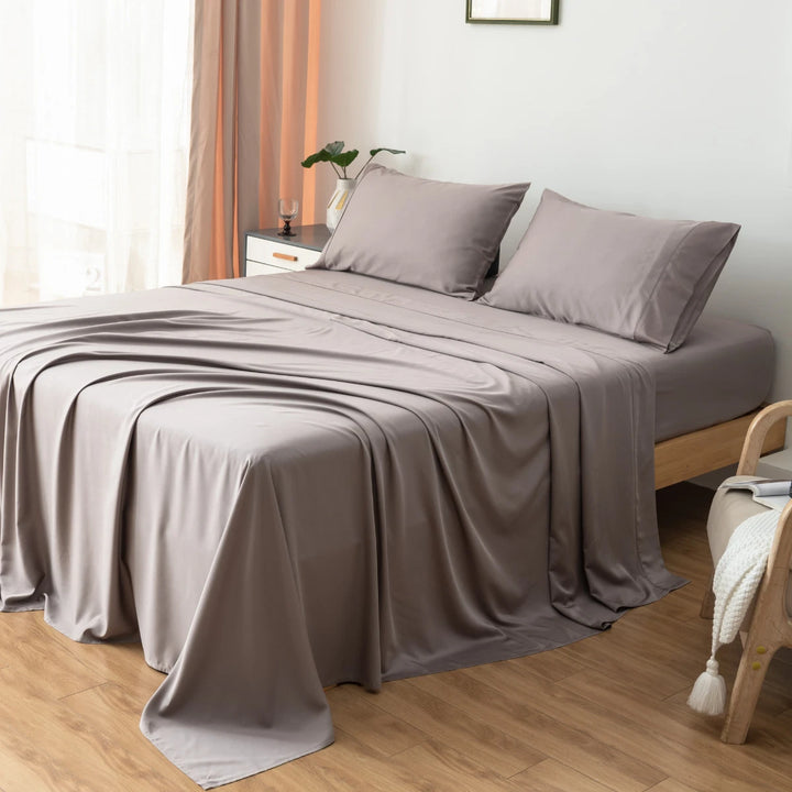 A neatly made bed with a smooth gray bedspread and Linenly's Bamboo Pillowcase Set - Stone Terrace in a bright room with wooden furniture and warm-toned curtains.