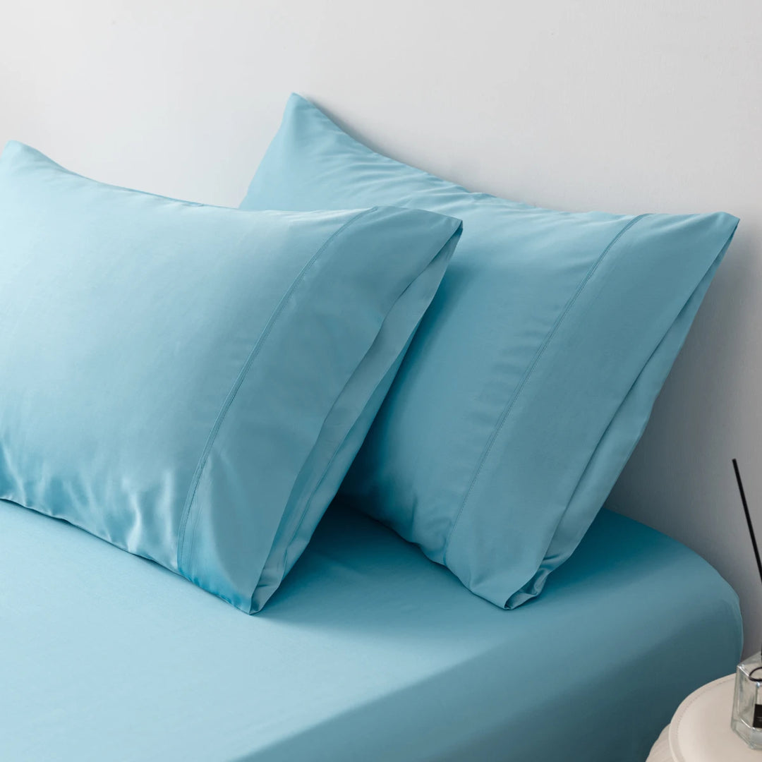 A neatly made bed with soft blue sheets and two Linenly Aqua Blue bamboo pillowcases against a white wall, offering a tranquil and cozy atmosphere for rest.