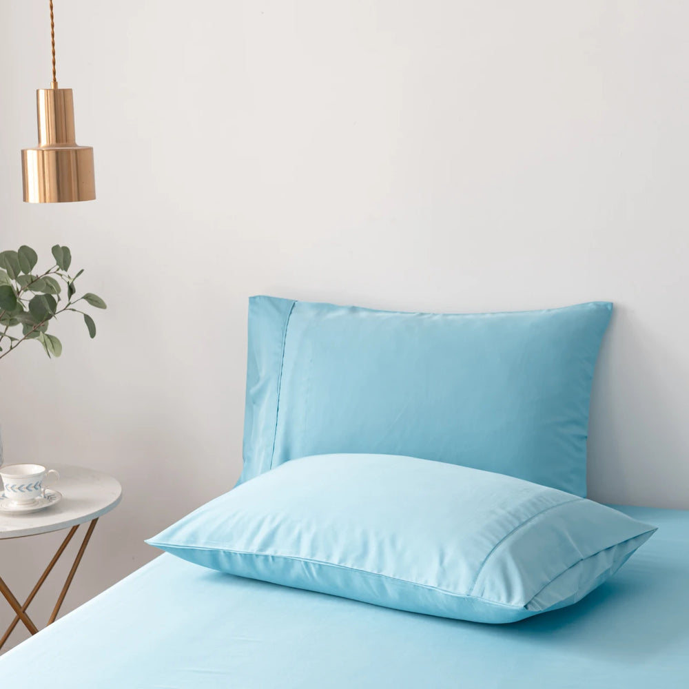 A neatly made bed with Linenly's Aqua Blue bamboo pillowcase set against a minimalist white wall, accompanied by a modern hanging lamp and a side table with a green plant.