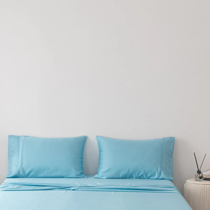 A minimalist bedroom with Linenly's Aqua Blue Bamboo Pillowcase Set on a neatly made bed and a small side table with decorative items, against a plain white wall, evoking a sense of calm and simplicity.