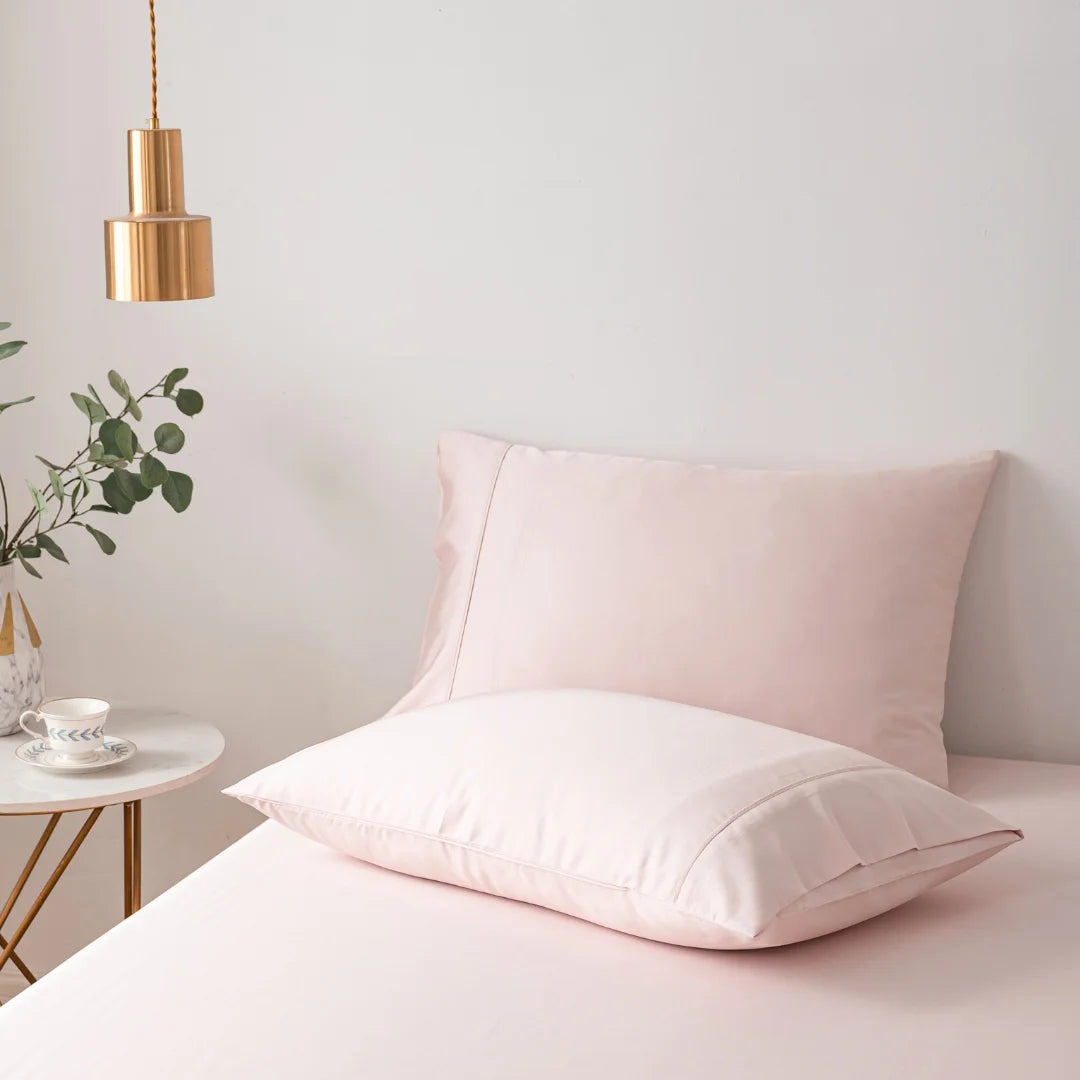 A serene and minimalist bedroom corner with a Linenly Bamboo Pillowcase Set in blush pink encasing a pillow on a matching bedsheet, complemented by a simple white nightstand holding a small plant and a pendant light.