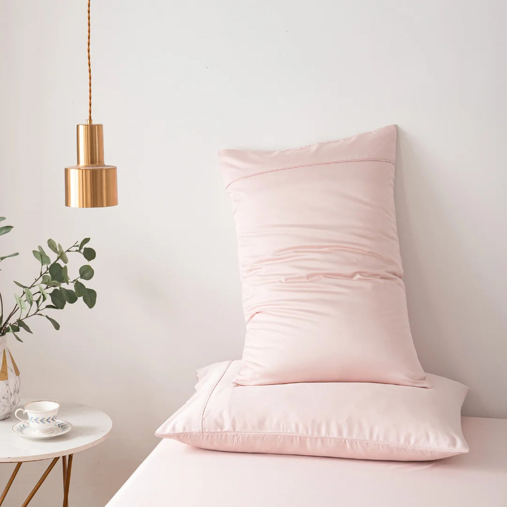 A cozy corner featuring a Linenly blush pink bamboo pillowcase set on a matching seat, with a modern gold pendant lamp overhead and a touch of greenery, creating a tranquil spot perfect for a quiet tea time.