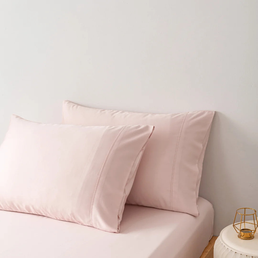A serene bedroom corner with soft pink pillows encased in Linenly organic bamboo pillowcases in Blush on a light sofa, next to a small bedside table with a minimalistic golden lamp.