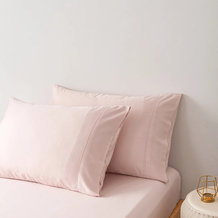 A serene bedroom corner with soft pink pillows encased in Linenly organic bamboo pillowcases in Blush on a light sofa, next to a small bedside table with a minimalistic golden lamp.