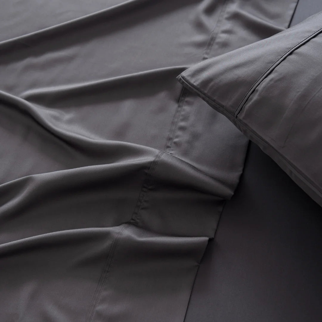 Smooth and sleek dark gray satin weave bed sheets and Linenly's Bamboo Pillowcase Set in Charcoal color arranged neatly, showcasing a comfortable and luxurious bedding set.