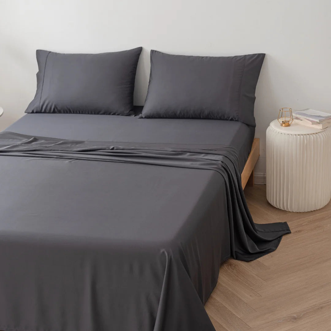 A neatly made bed with a dark gray bedsheet and Linenly Bamboo Pillowcase Set - Charcoal, accompanied by a bedside table and a lamp, in a room with light wooden flooring.