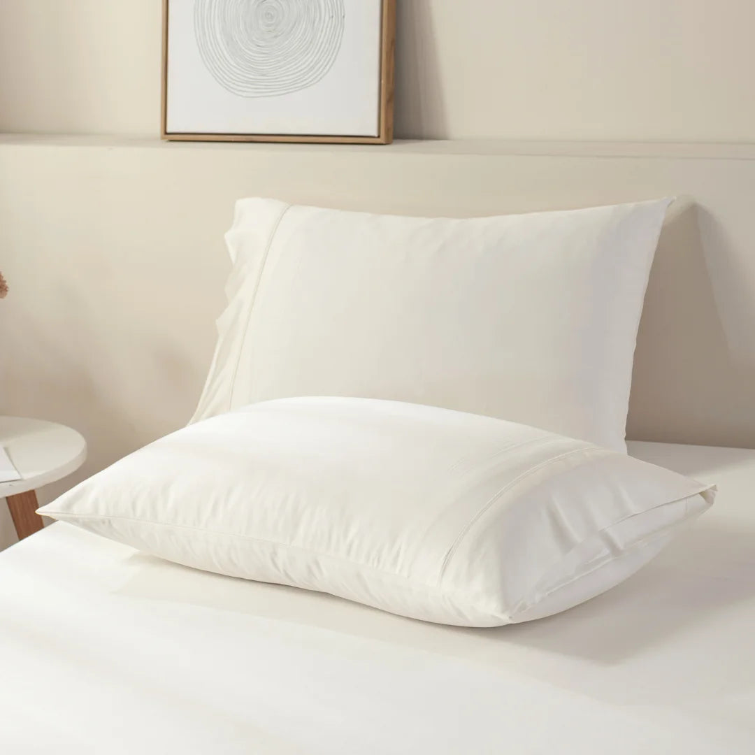 A pair of clean, white pillows with Linenly organic bamboo pillowcases in Ivory on a neatly made bed with a neutral-toned backdrop and minimalistic decor.
