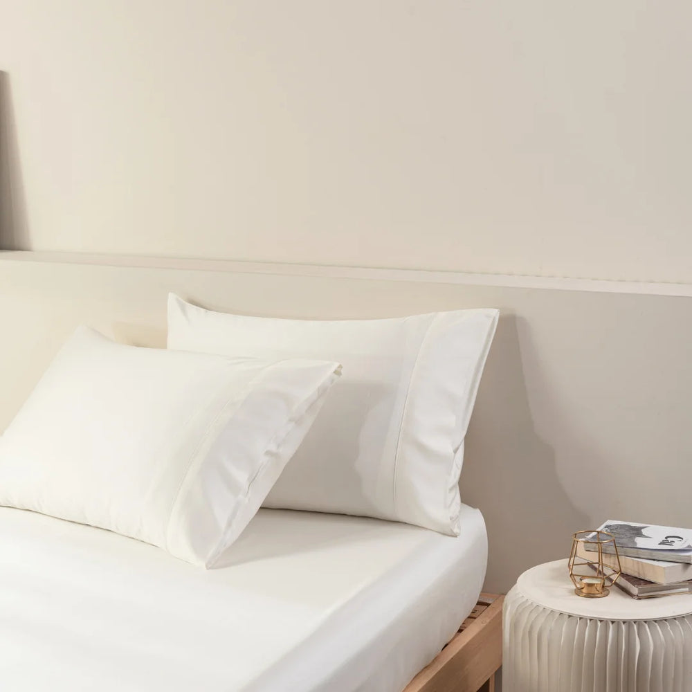 A neatly made bed with crisp white linens and fluffy pillows encased in Linenly organic bamboo pillowcases, accompanied by a small bedside table topped with a stack of books and glasses.