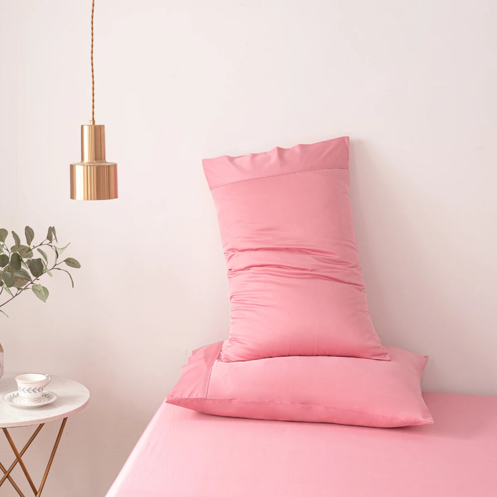 A neatly arranged pink pillow with Linenly bamboo pillowcases on a matching pink bedsheet, with a minimalist decor including a hanging copper lamp and a small plant on a bedside table in a room with light-colored walls