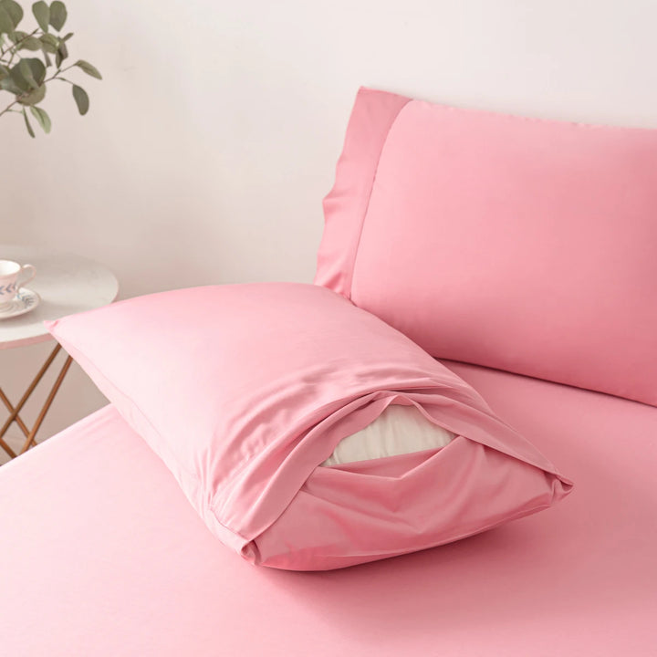 A neatly made bed with soft pink sheets and a Linenly Bamboo Pillowcase Set - Light Rose pillow with an open pillowcase, set against a minimalist background.