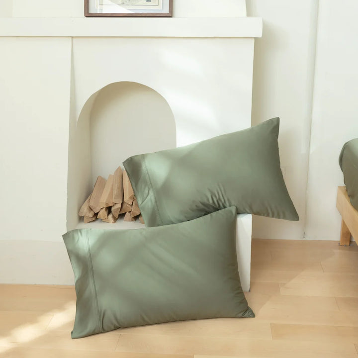 Two Linenly Bamboo Pillowcase Set - Moss, casually set on a wooden floor near a fireplace with logs.