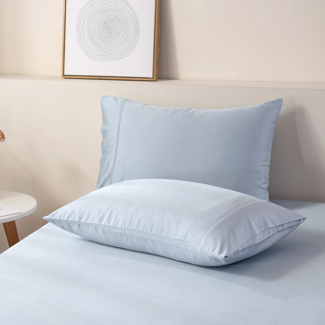 Two fluffy pillows encased in a Linenly organic Bamboo Pillowcase Set in Pale Blue sit atop a neatly made bed with light blue linens against a soft-hued background, creating a serene and inviting atmosphere in a modern bedroom.
