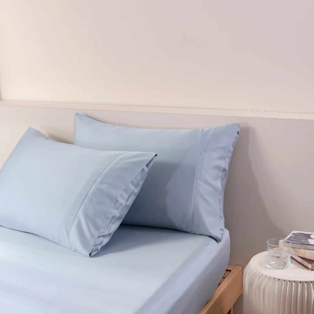 Crisp blue bed sheets and a Linenly Bamboo Pillowcase Set in pale blue satin weave on a neatly made bed, inviting a peaceful night's sleep.